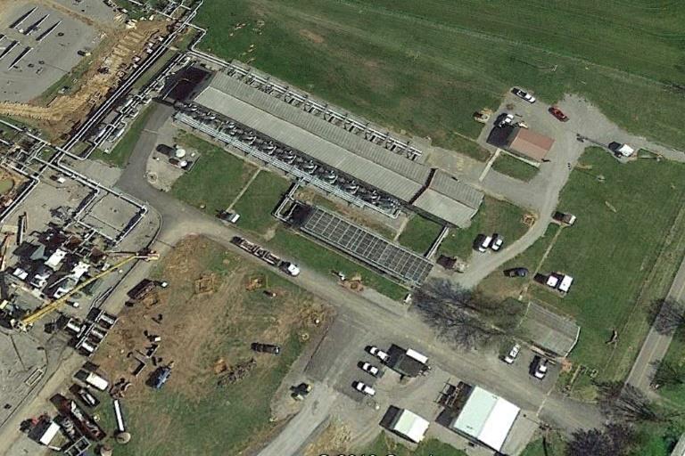 Aerial view of an industrial site featuring a large building, surrounding green space, and several parked vehicles.