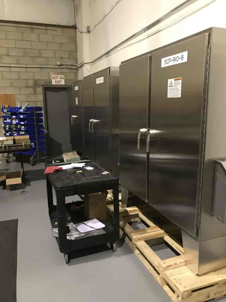Industrial control panels in a warehouse with storage shelves in the background.