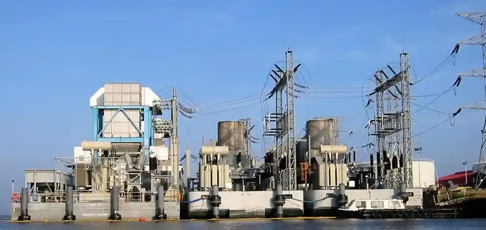Industrial barge structure on calm water with transformers and power cables under a clear sky.