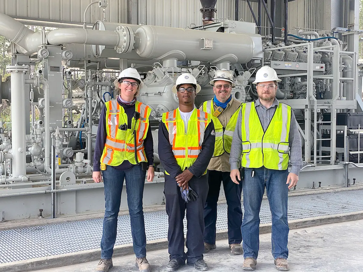 Group of industrial workers in safety vests and hard hats standing in front of large machinery at a worksite.